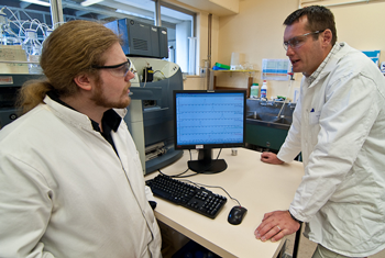 Two male scientists wearing labcoats and safety glasses discussing results over a desk with a computer in the background.