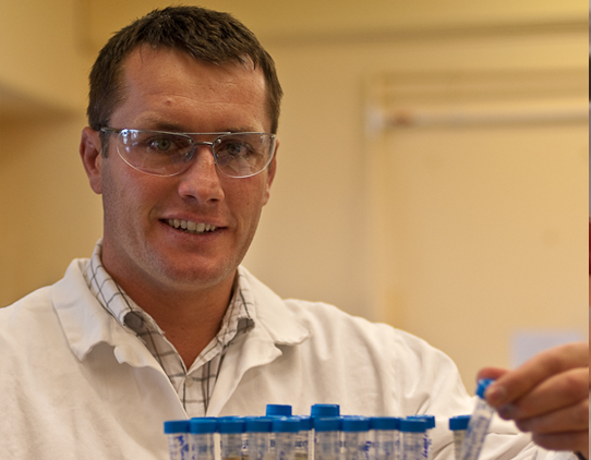 A scientist wearing a lab coat and safety glasses holds a rack of test tubes. They contain clear, yellow liquid.