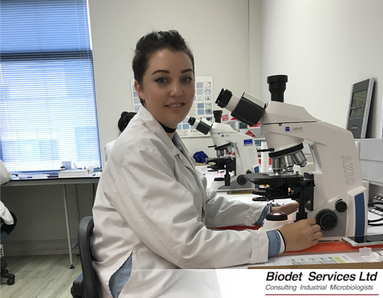 A scientist wearing a labcoat and safety glasses is sitting at a microscope. She is smiling.