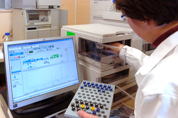 A scientist wearing a labcoat places vials into an instrument. A computer monitor is in the foreground.